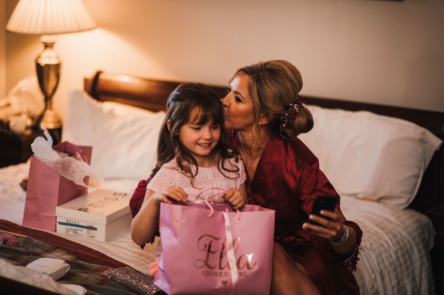 Two people share an intimate moment while opening a pink gift bag on a bed in warm lighting.
