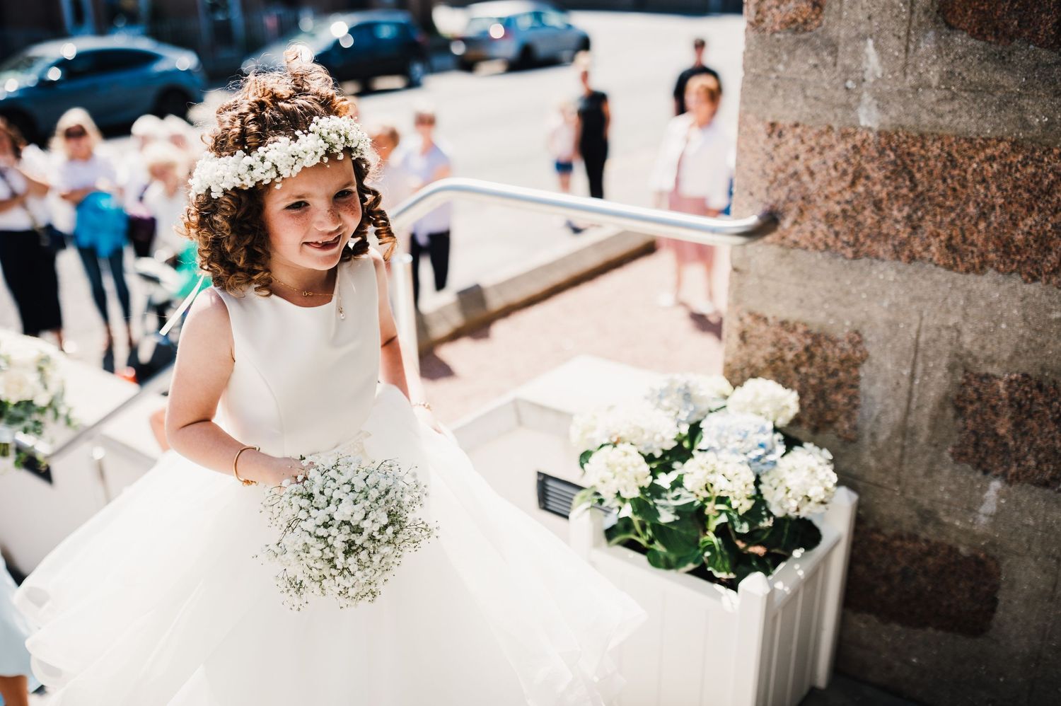 Young flower girl in white dress holds baby's breath bouquet outside church.