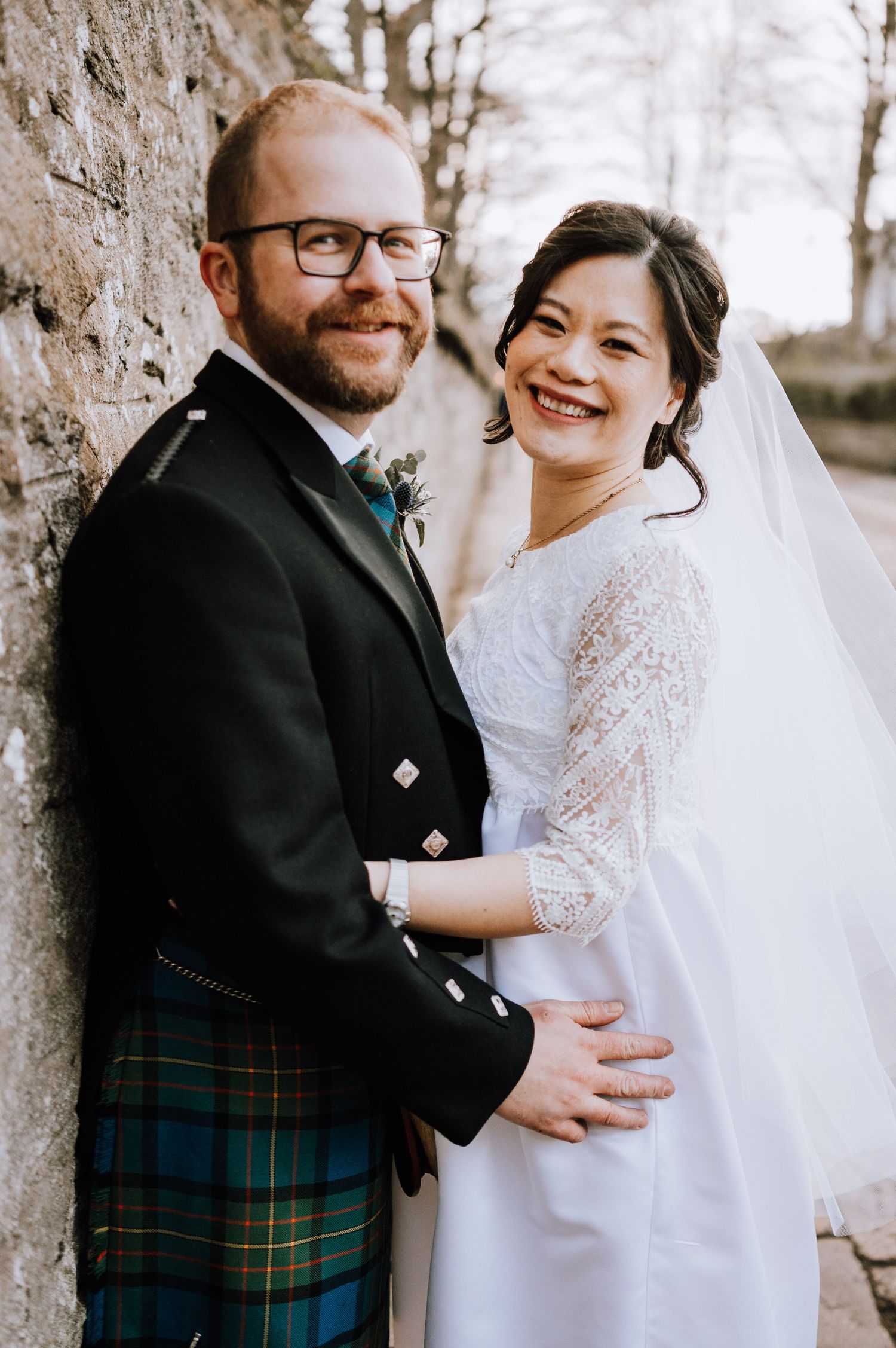 Happy wedding couple in Scottish and white wedding attire pose against stone wall.