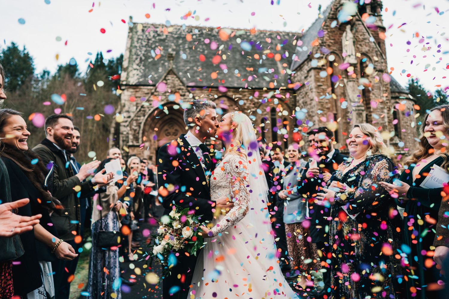 Colorful confetti fills the air as wedding guests celebrate outside a stone church.