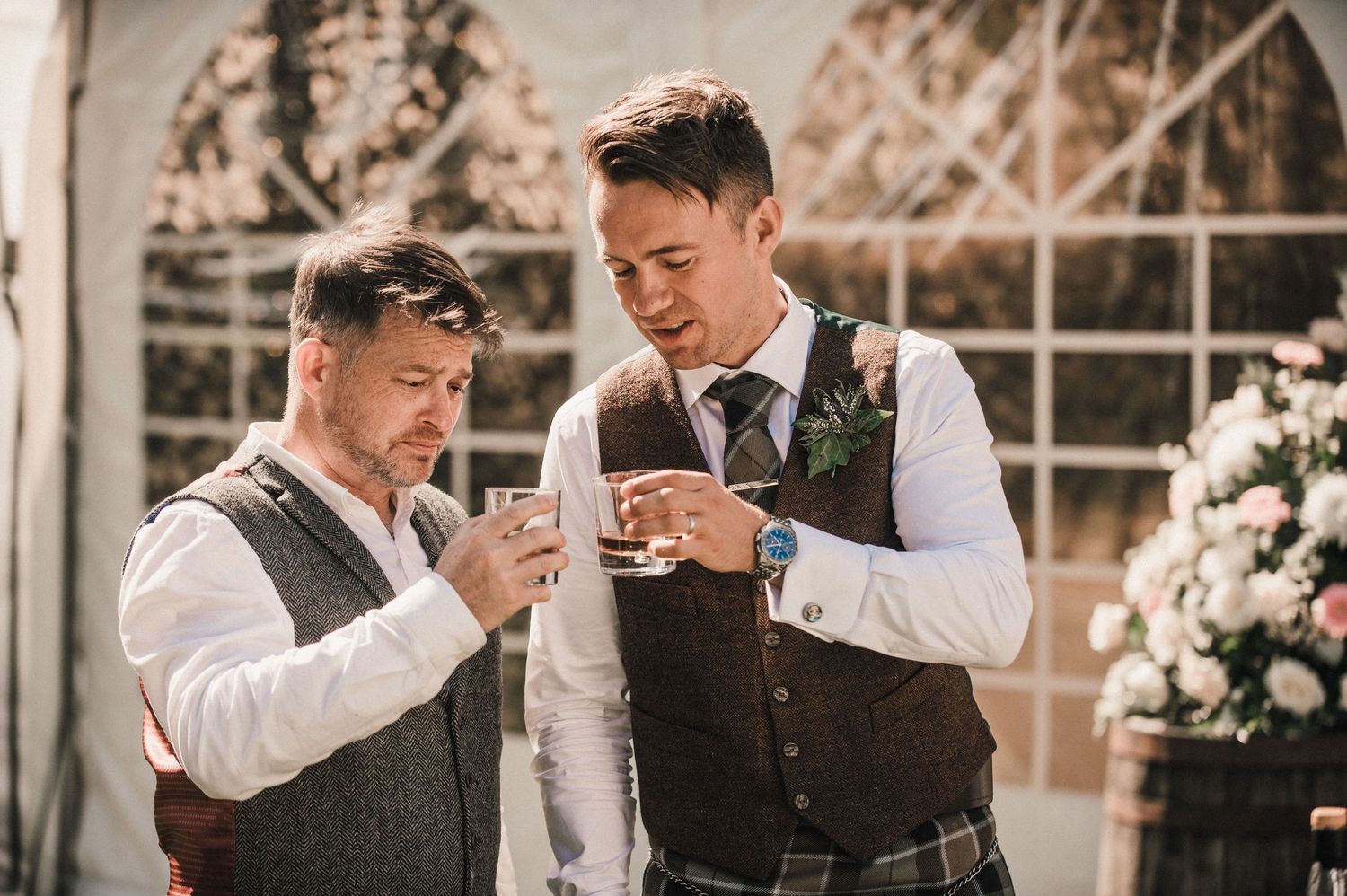 Two wedding guests sharing drinks and laughter in a sunlit greenhouse.