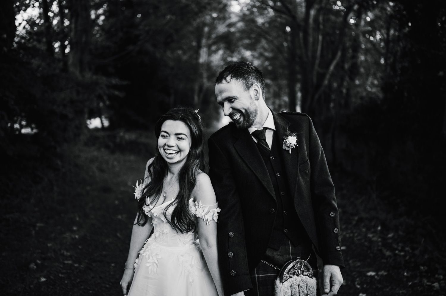 A black and white candid photo of a laughing couple in wedding attire surrounded by trees.
