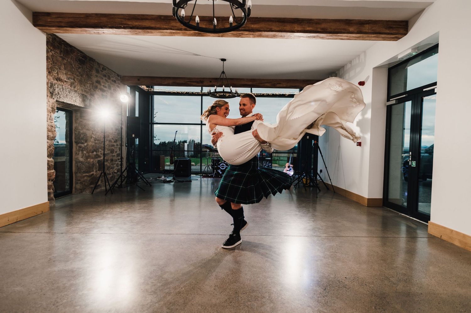 Groom in Scottish kilt lifts bride dramatically during their wedding dance in a modern venue.