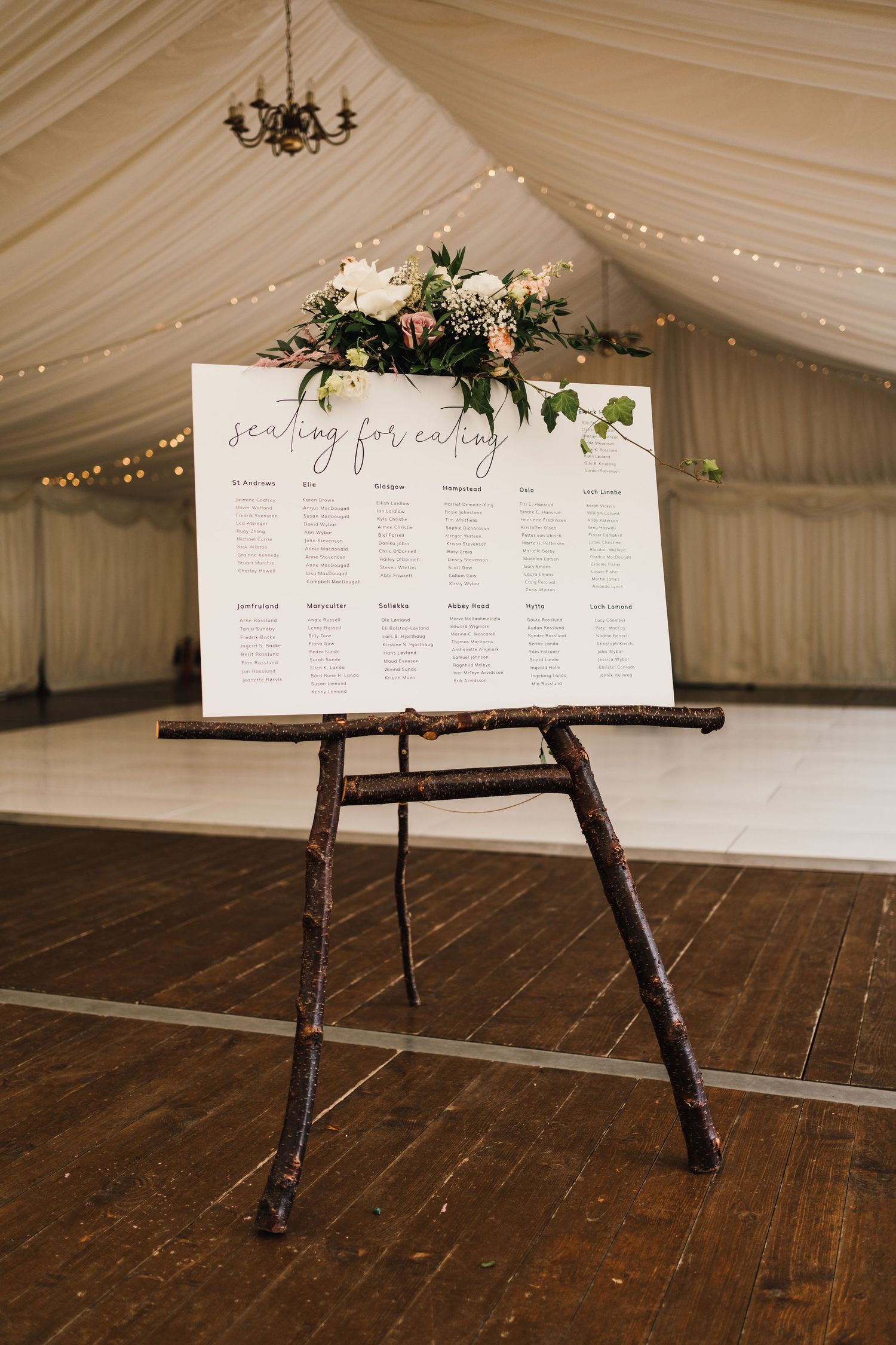 Wedding seating chart displayed on wooden easel with floral arrangement and fairy lights in marquee setting.