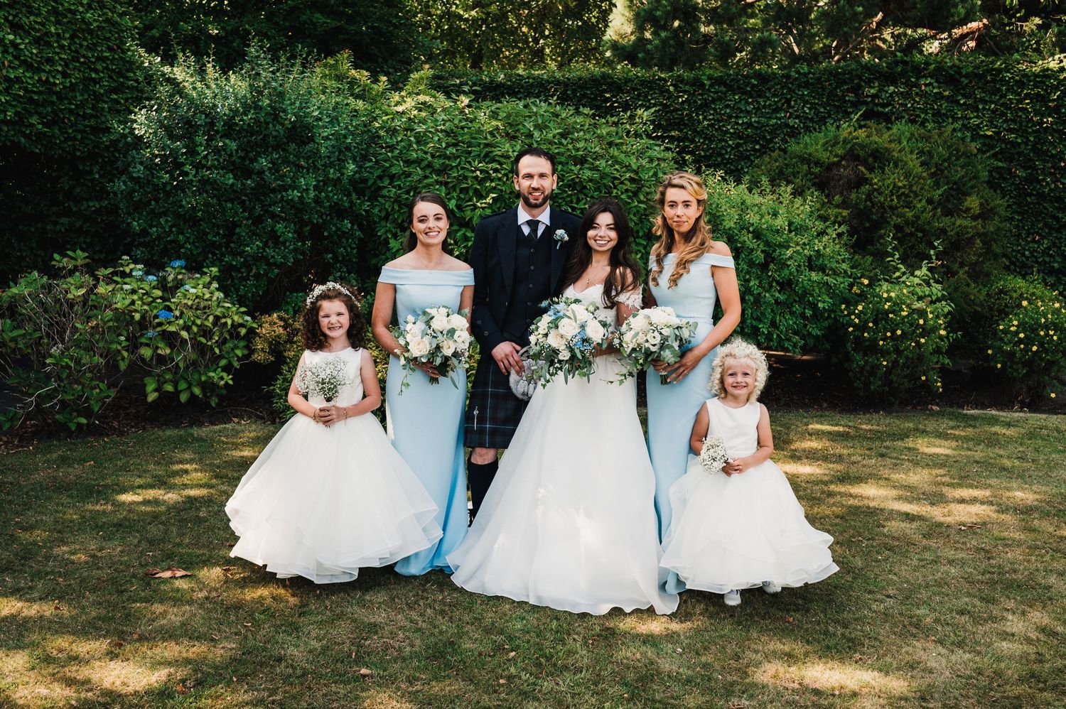 Wedding party poses in garden setting with mix of formal dresses and Scottish attire.