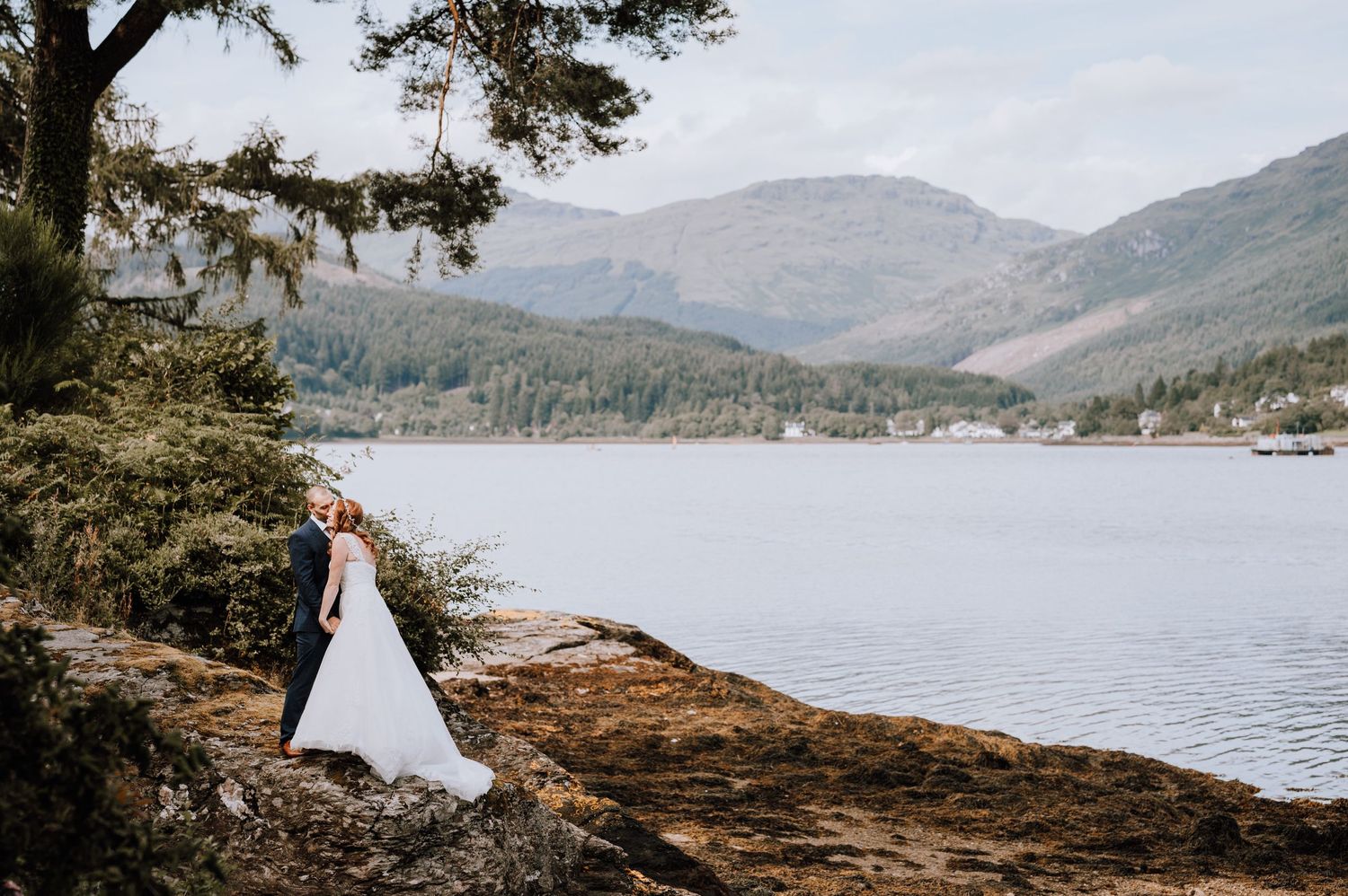Wedding couple poses on shoreline with stunning mountain lake vista in background.