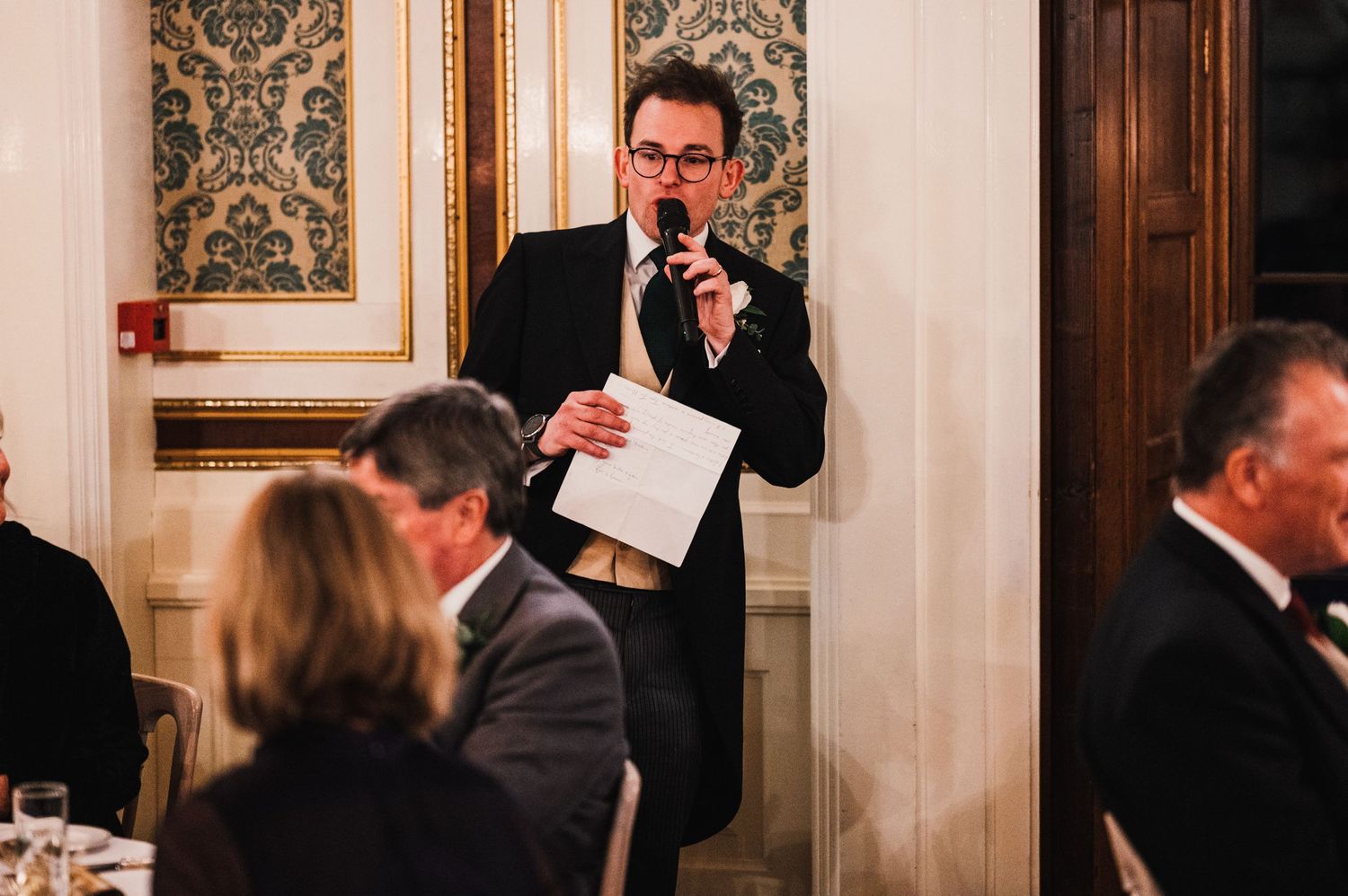 Someone gives a speech at an elegant dinner reception with ornate wallpaper and decor.