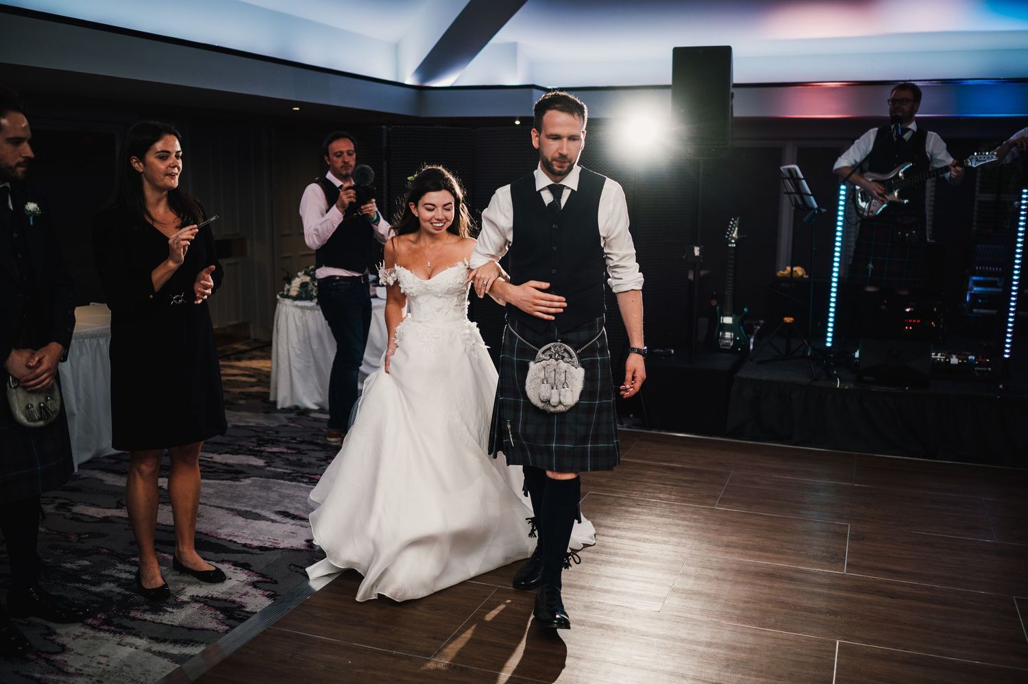 Wedding couple walks across dance floor during reception while guests look on.