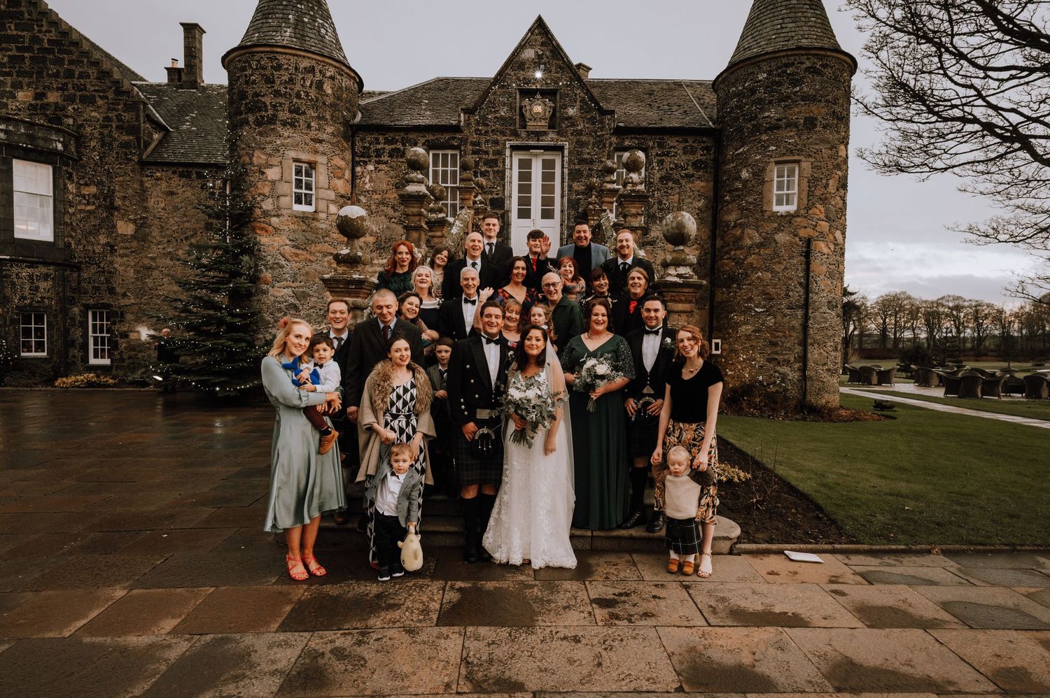 Large wedding party group photo outside historic Scottish castle on overcast day.