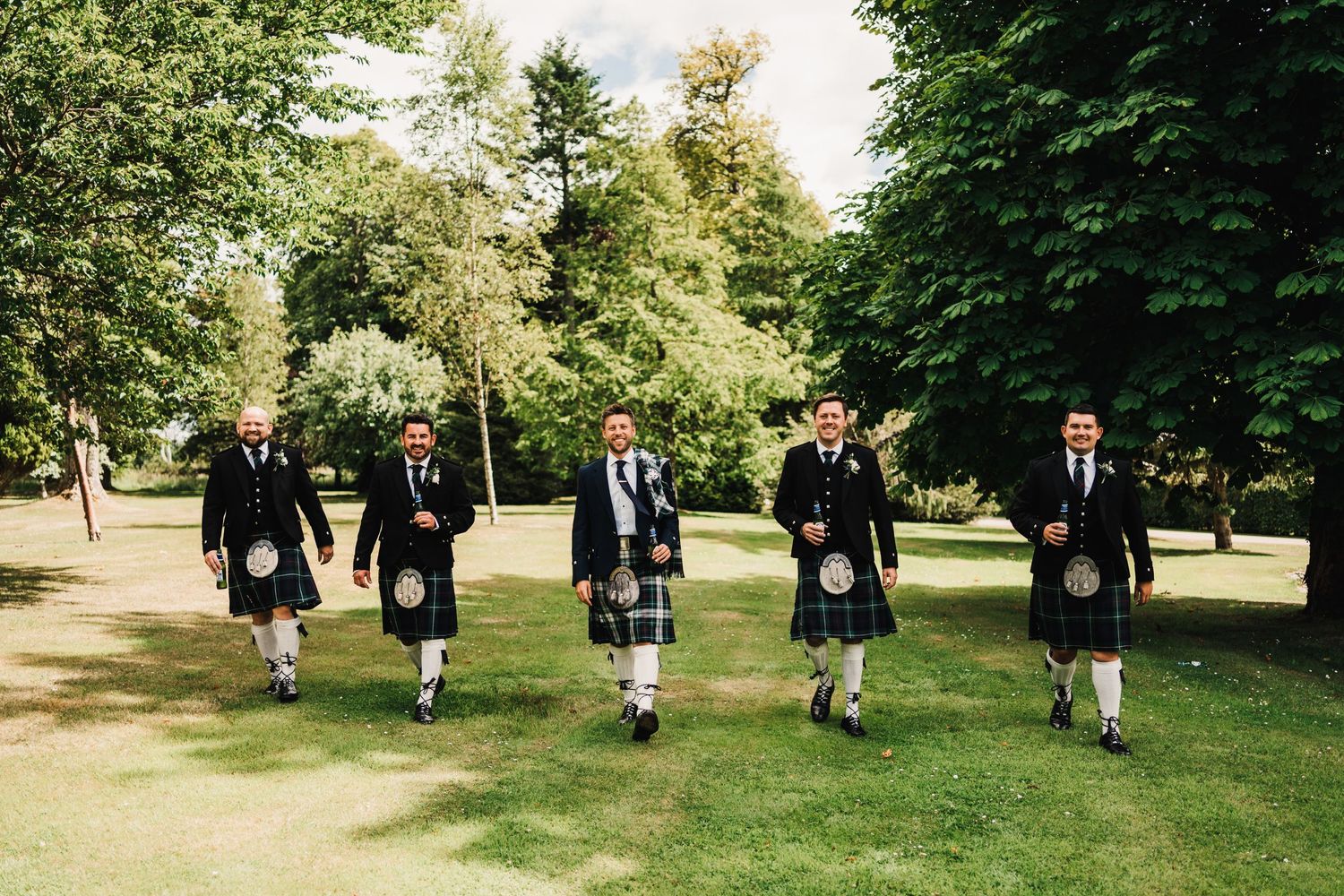 Five groomsmen in traditional Scottish kilts walk across sunny garden lawn.