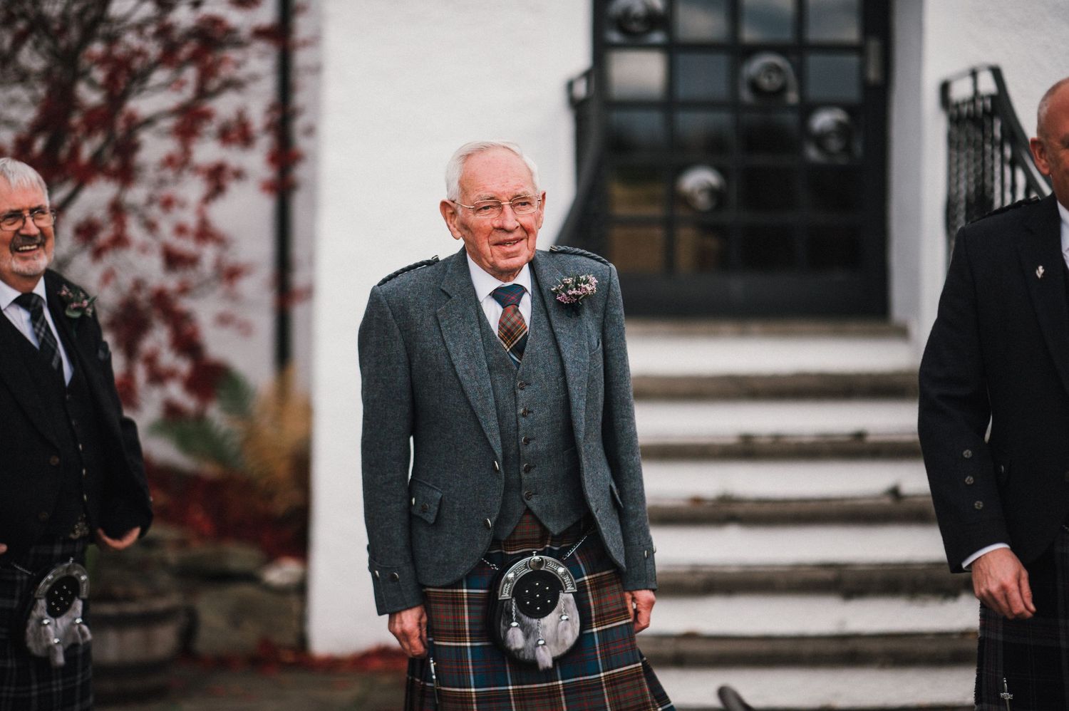 Three people in traditional Scottish formal wear stand on white steps outside a building.
