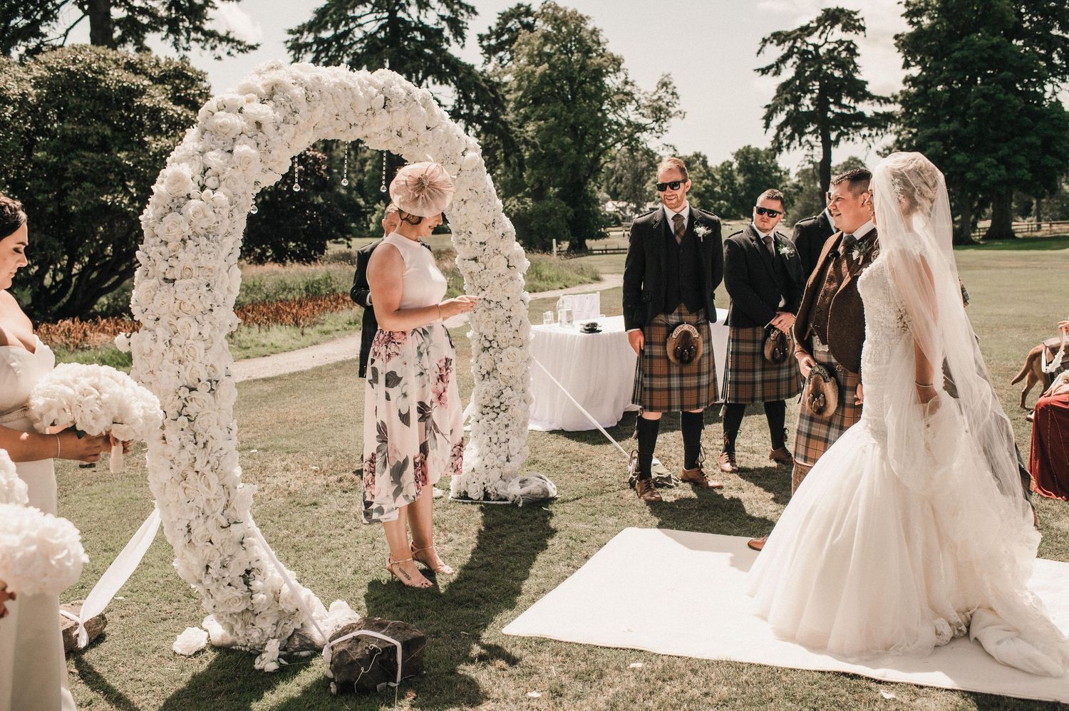 Wedding ceremony under a white floral arch with Scottish-themed attire and decorations.