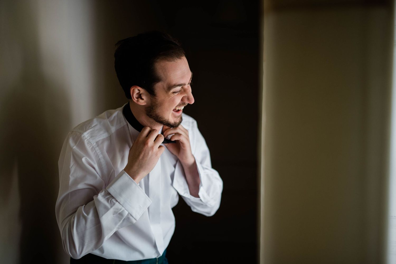 A formally dressed person adjusting their collar in dramatic lighting near a window.