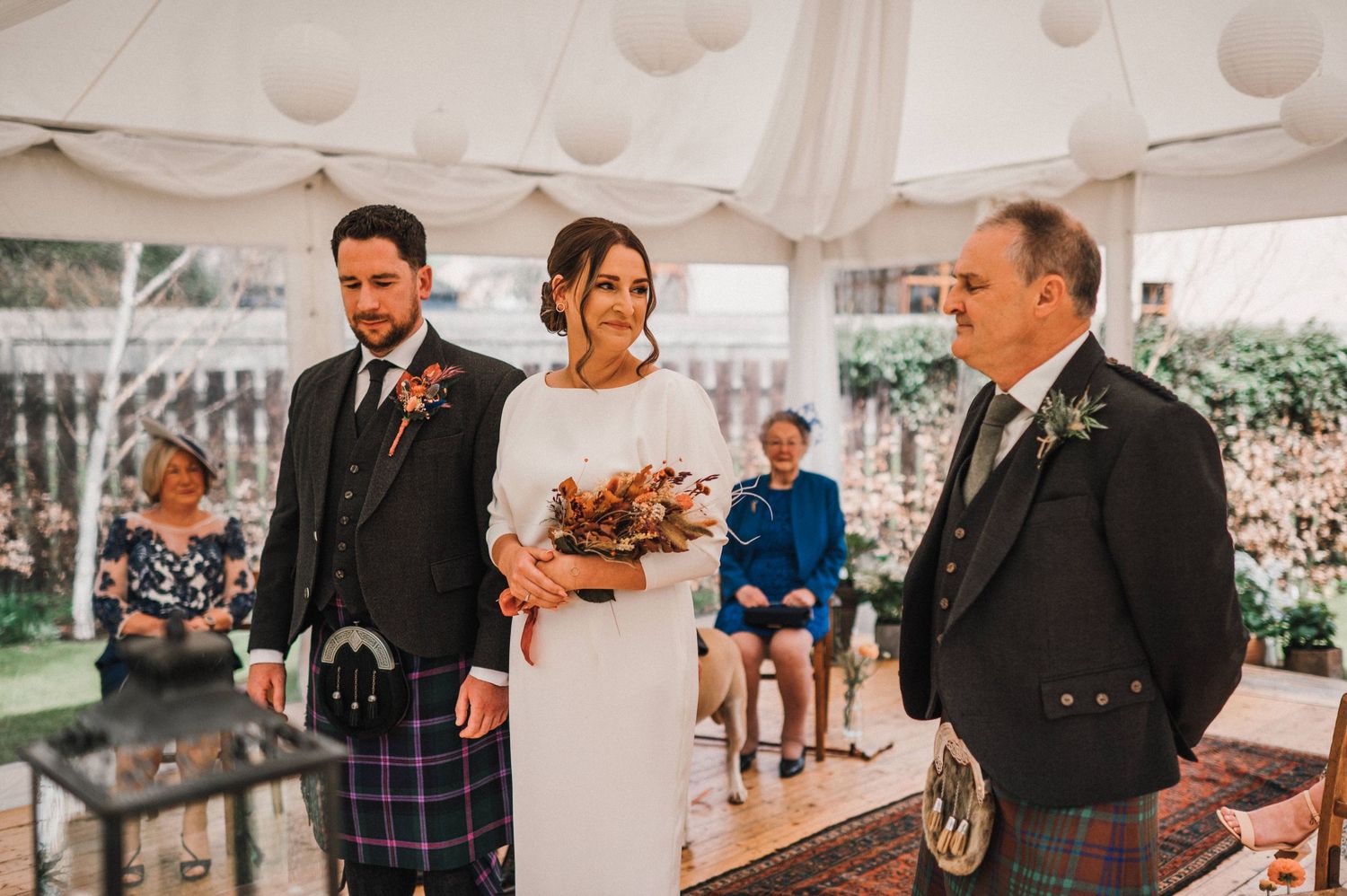 A wedding ceremony in progress under a white tent with guests seated and Scottish attire on display.