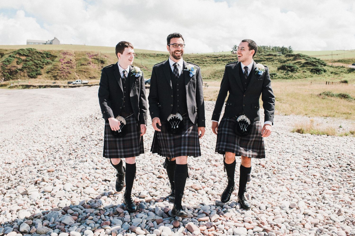 Three groomsmen in matching kilts walk together on a rocky Scottish beach.