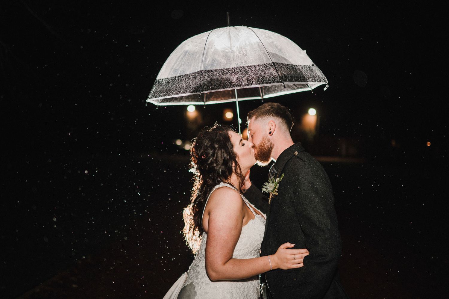 Couple embraces under clear umbrella during nighttime wedding photoshoot.