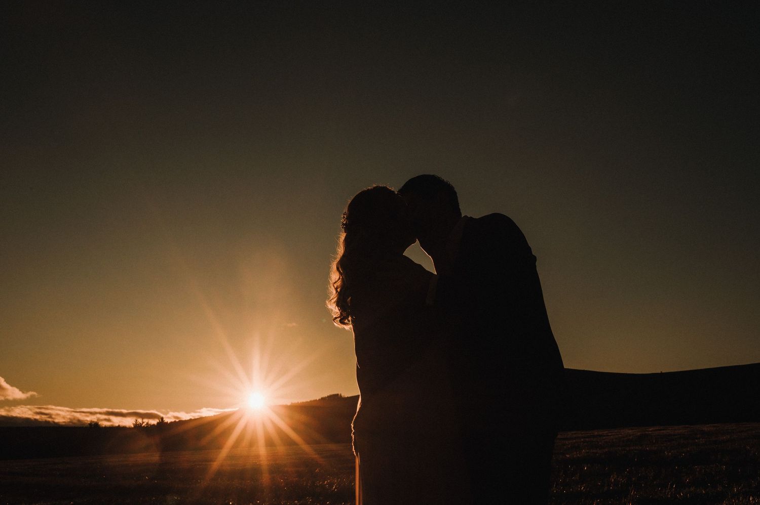 Silhouetted figures embrace as the sun sets behind rolling hills.