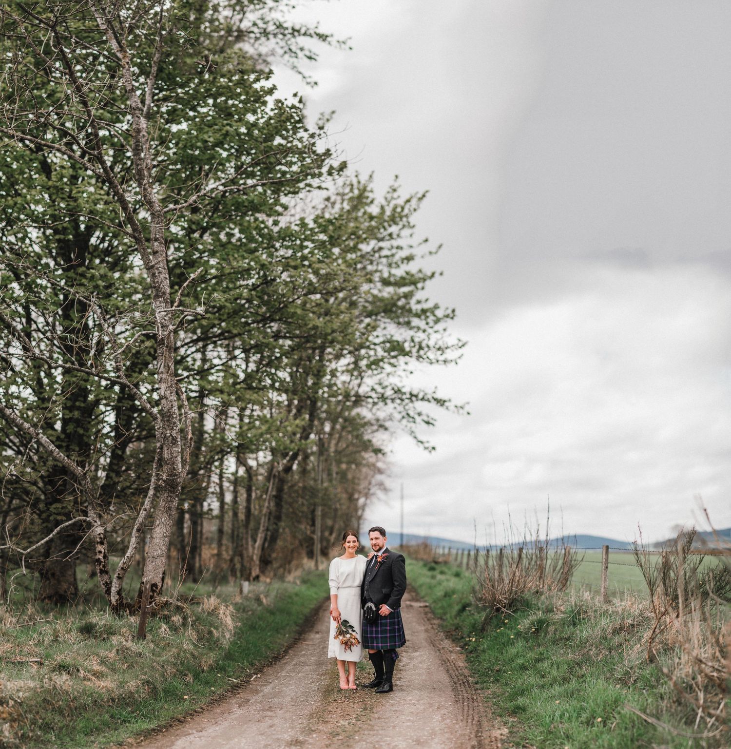Scenic wedding photo on a tree-lined rural path with rolling hills in the background.