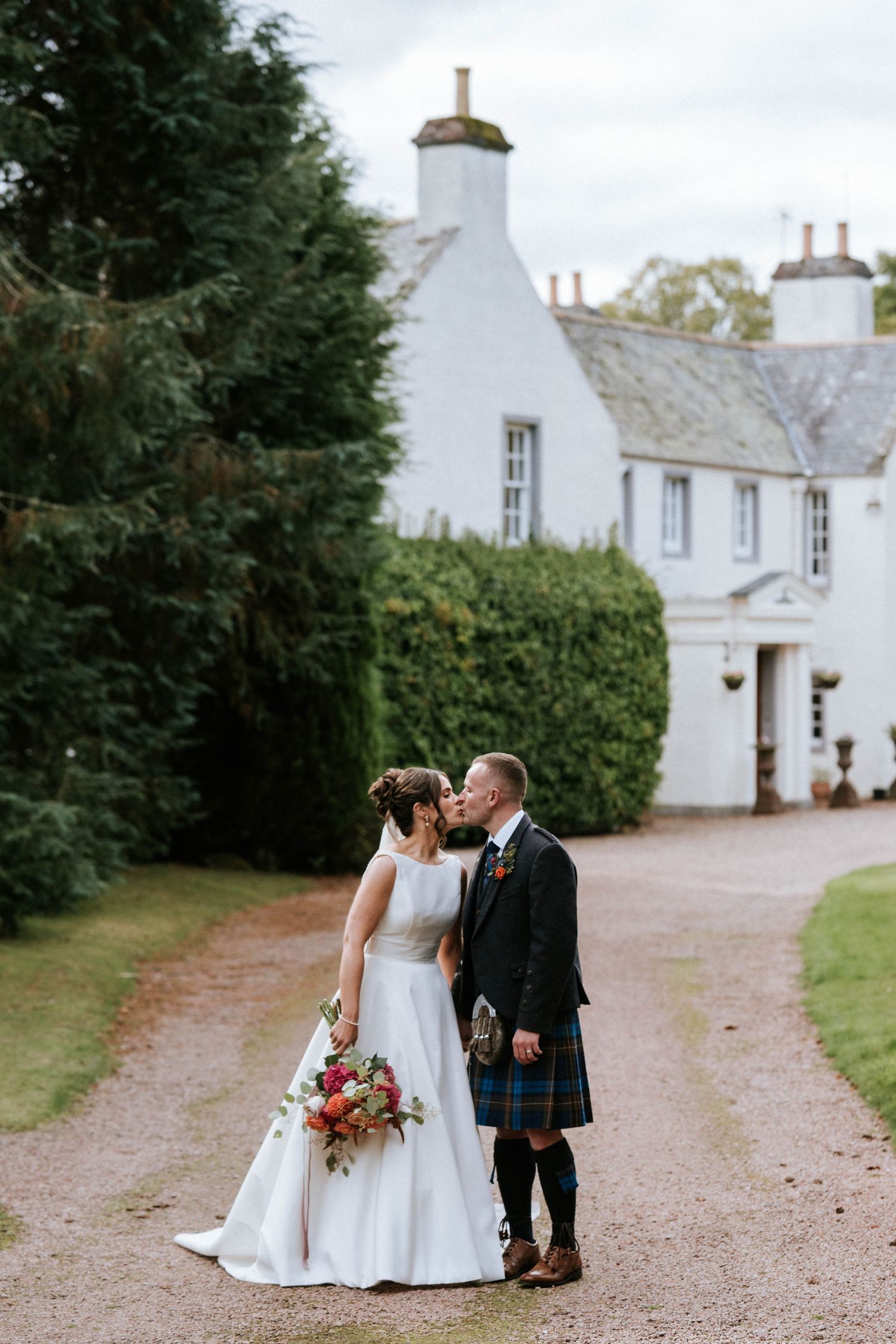 Wedding couple walking along gravel path beside traditional white Scottish estate buildings.