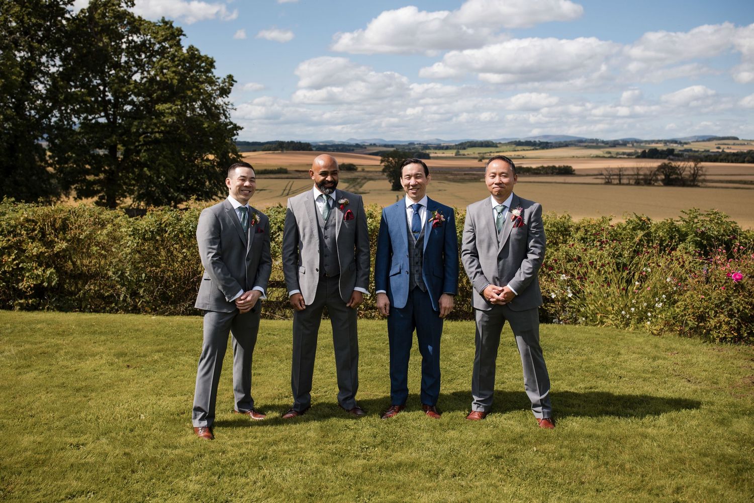 Wedding party members pose casually on a grassy hillside with scenic farmland and blue skies in the background.