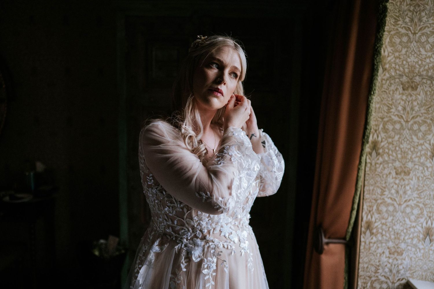 A lace wedding dress hangs elegantly against a textured wallpaper background in natural light.