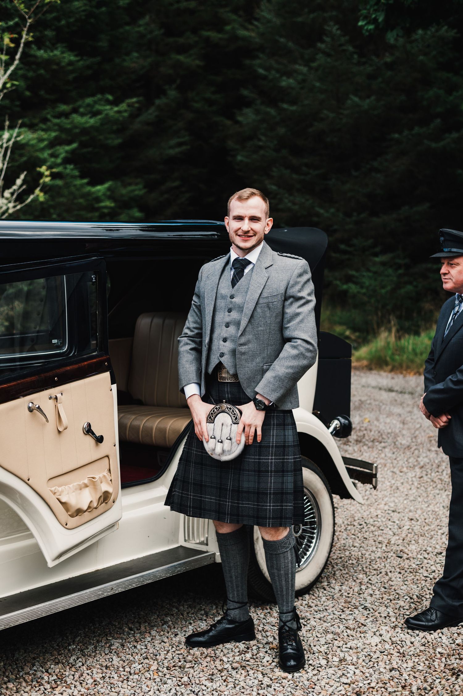 Wedding party members in traditional Scottish kilts and gray suits stand beside a classic white vintage car.