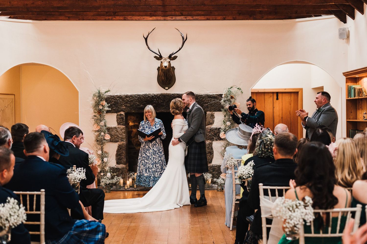 Wedding ceremony in progress at a rustic indoor venue with white chairs and wooden beam ceiling.