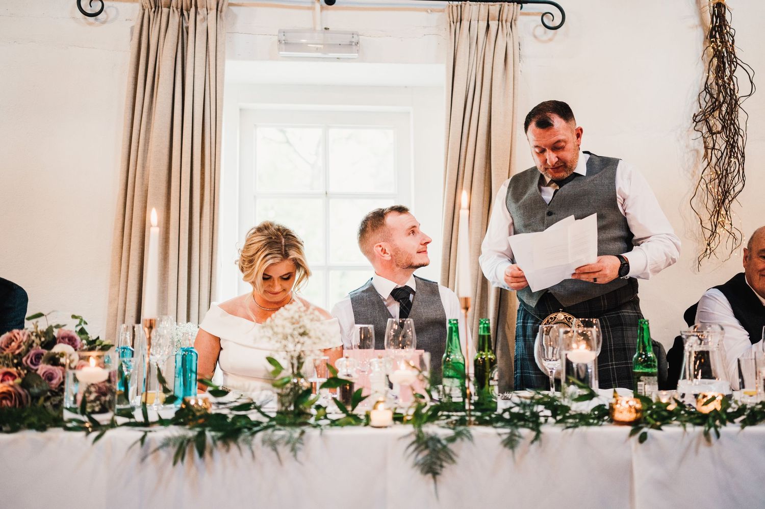 Reception scene with guests at head table decorated with greenery and blue bottles during wedding speeches.