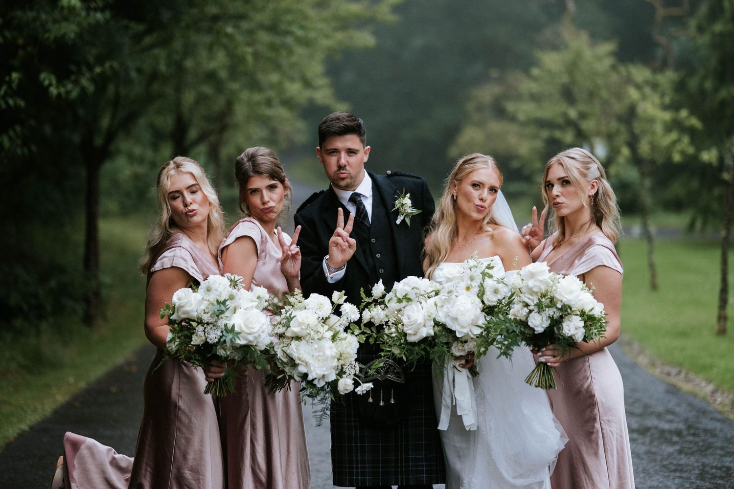 Wedding party poses outdoors with bridesmaids in blush dresses holding white floral bouquets.
