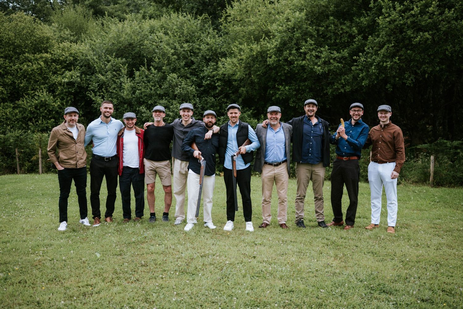 Large wedding party group photo outdoors with groomsmen and bridesmaids lined up on grass.