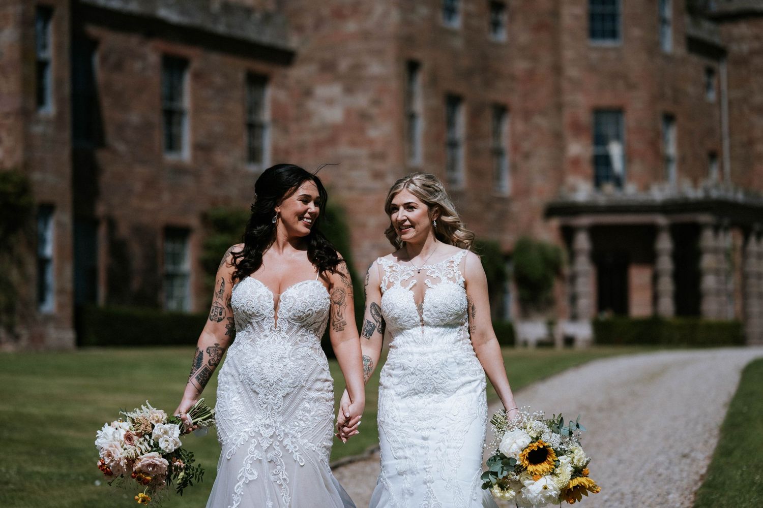 Two brides in fitted lace wedding dresses holding bouquets on castle grounds.