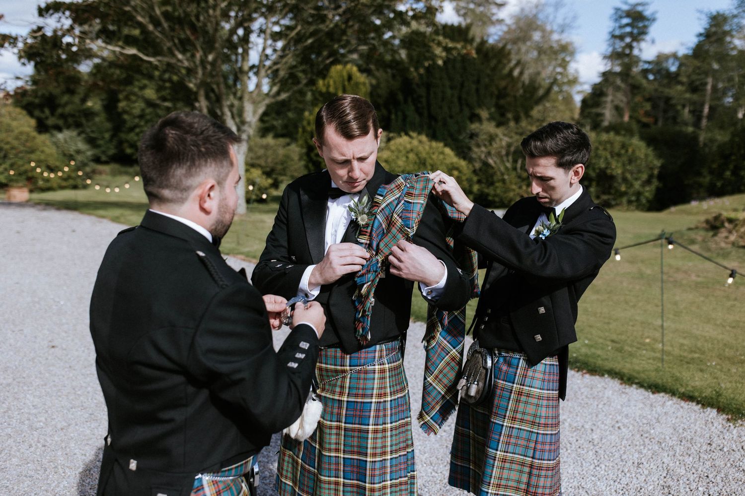 Groomsmen in kilts and formal attire walking outdoors on a sunny day.