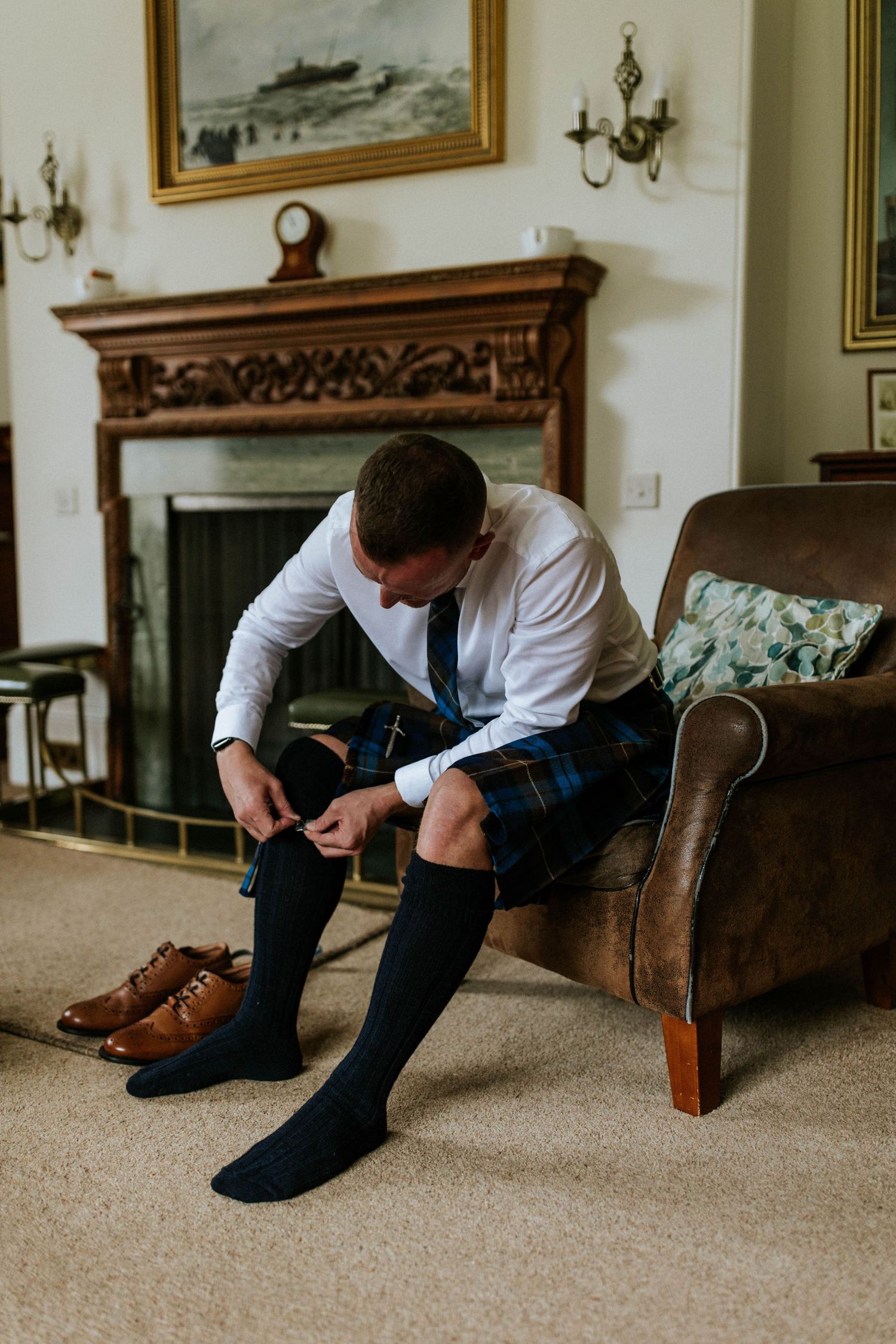 Person putting on brown leather dress shoes while wearing traditional Scottish kilt.