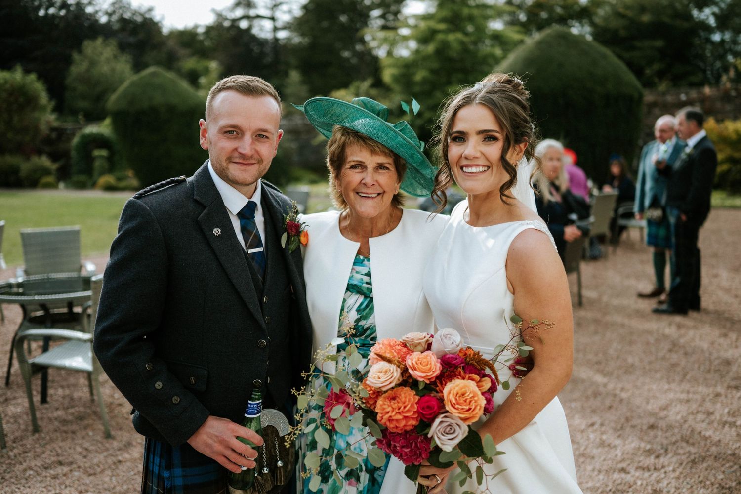 A wedding party poses outdoors with colorful bouquets and formal attire including a traditional Scottish kilt.