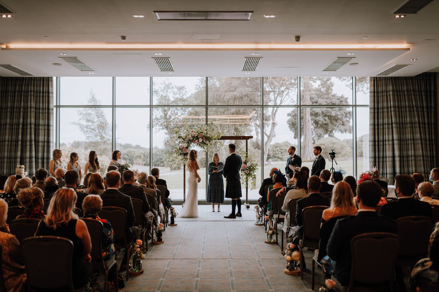 Modern wedding ceremony venue with floor-to-ceiling windows and guests seated in rows.