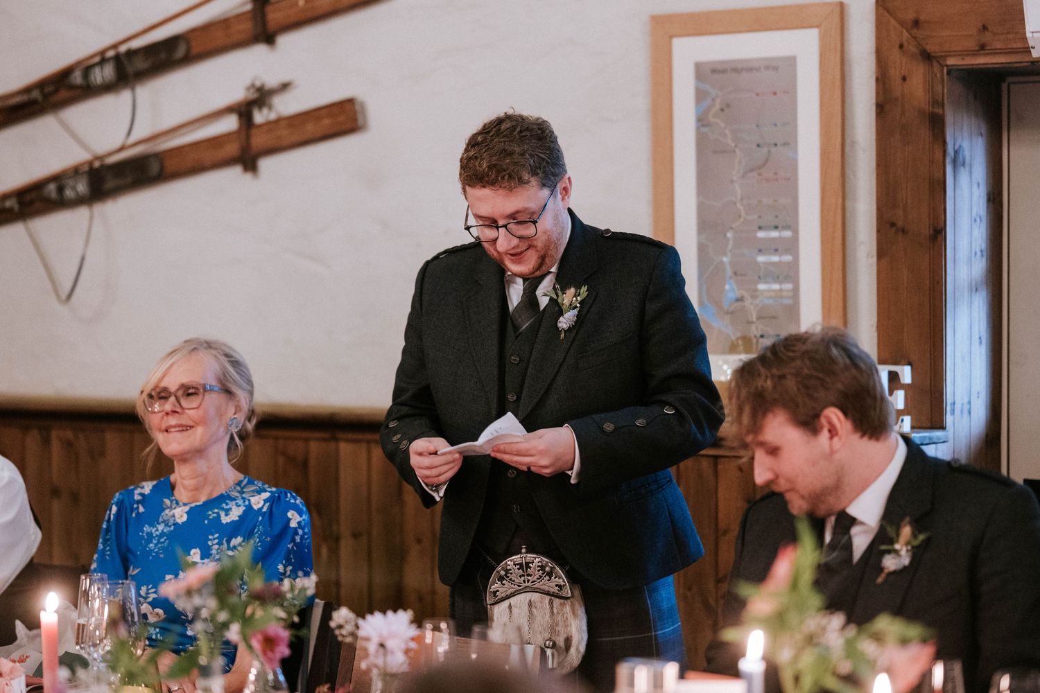 A wedding reception dinner scene with guests at a table while someone delivers a speech.