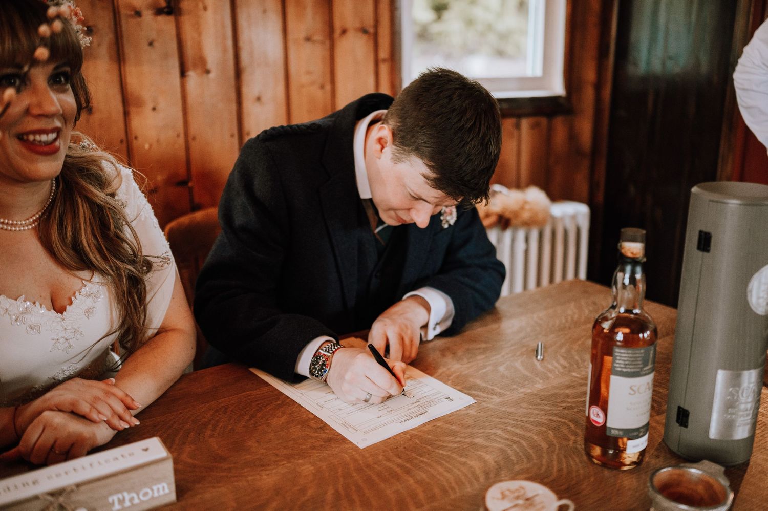 Signing wedding documents at a wooden table with a bottle of spirits nearby.