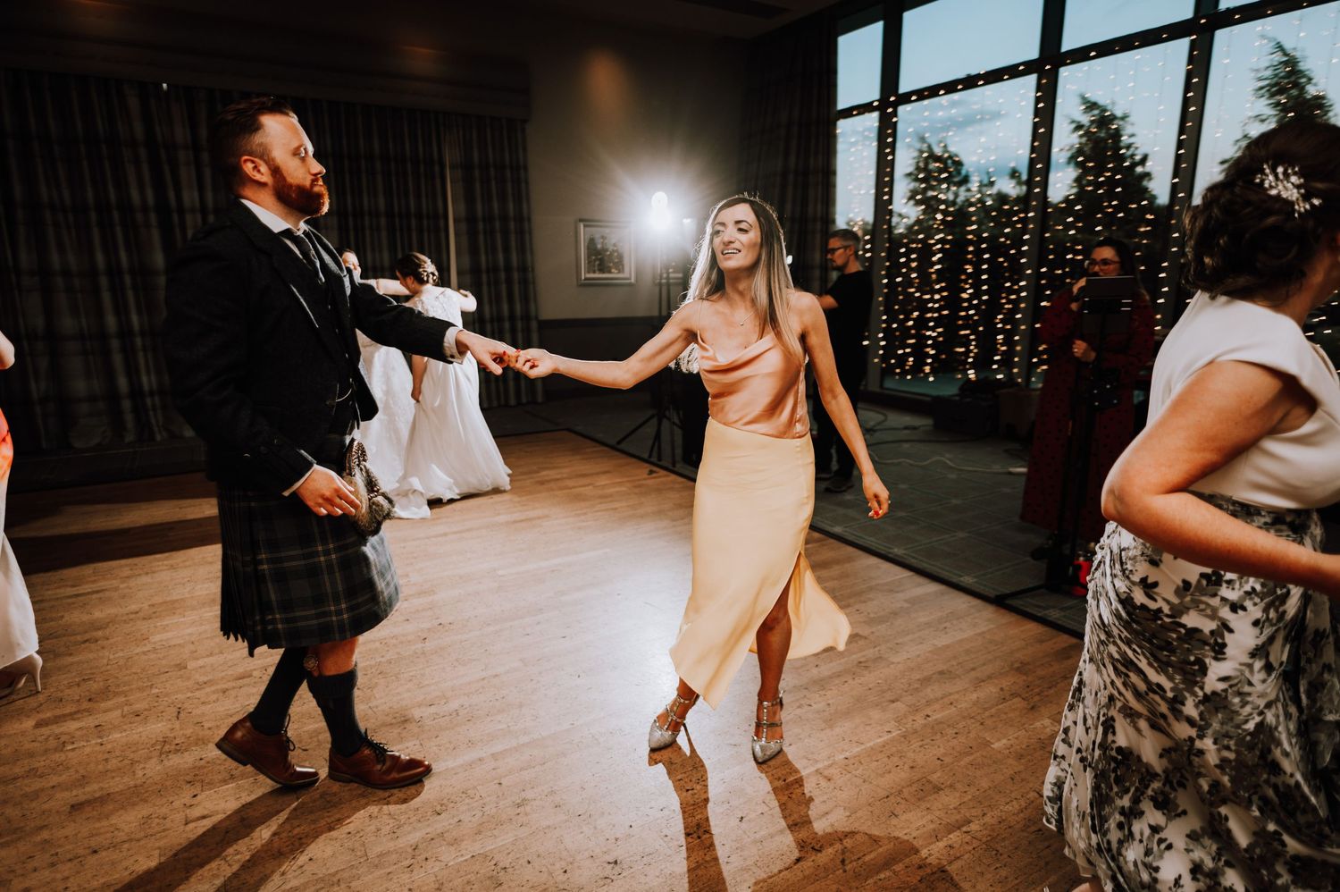 Wedding guests dance on a wooden floor with string lights visible through large windows.