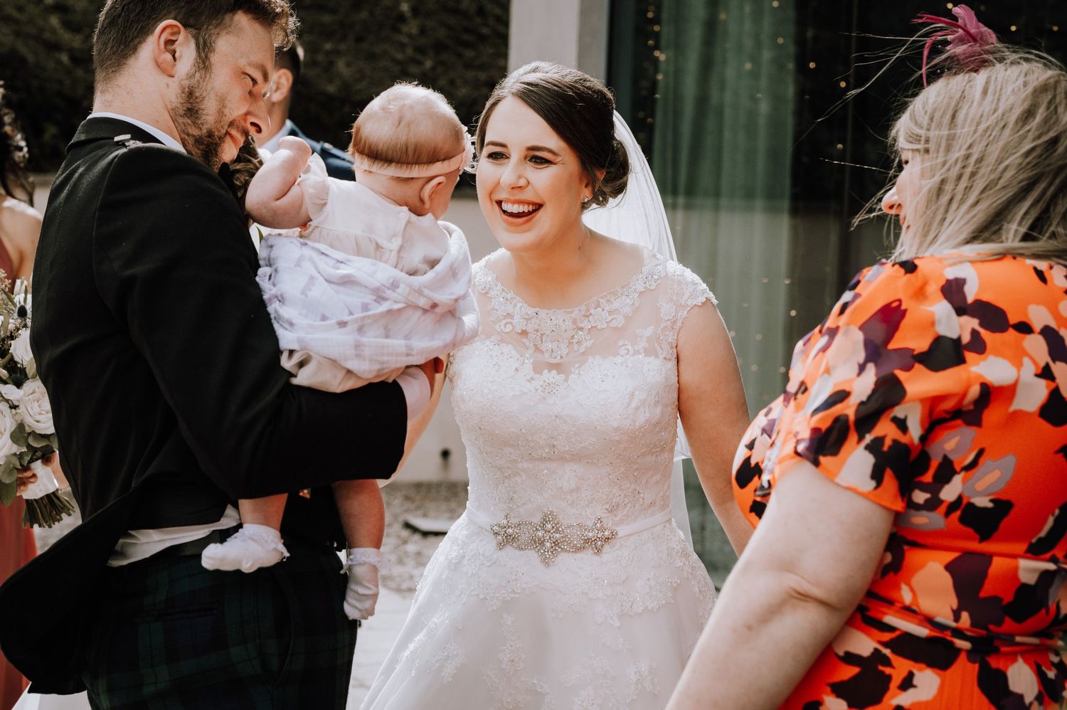 A joyful moment at a wedding with a couple holding a baby wearing white while other guests look on.