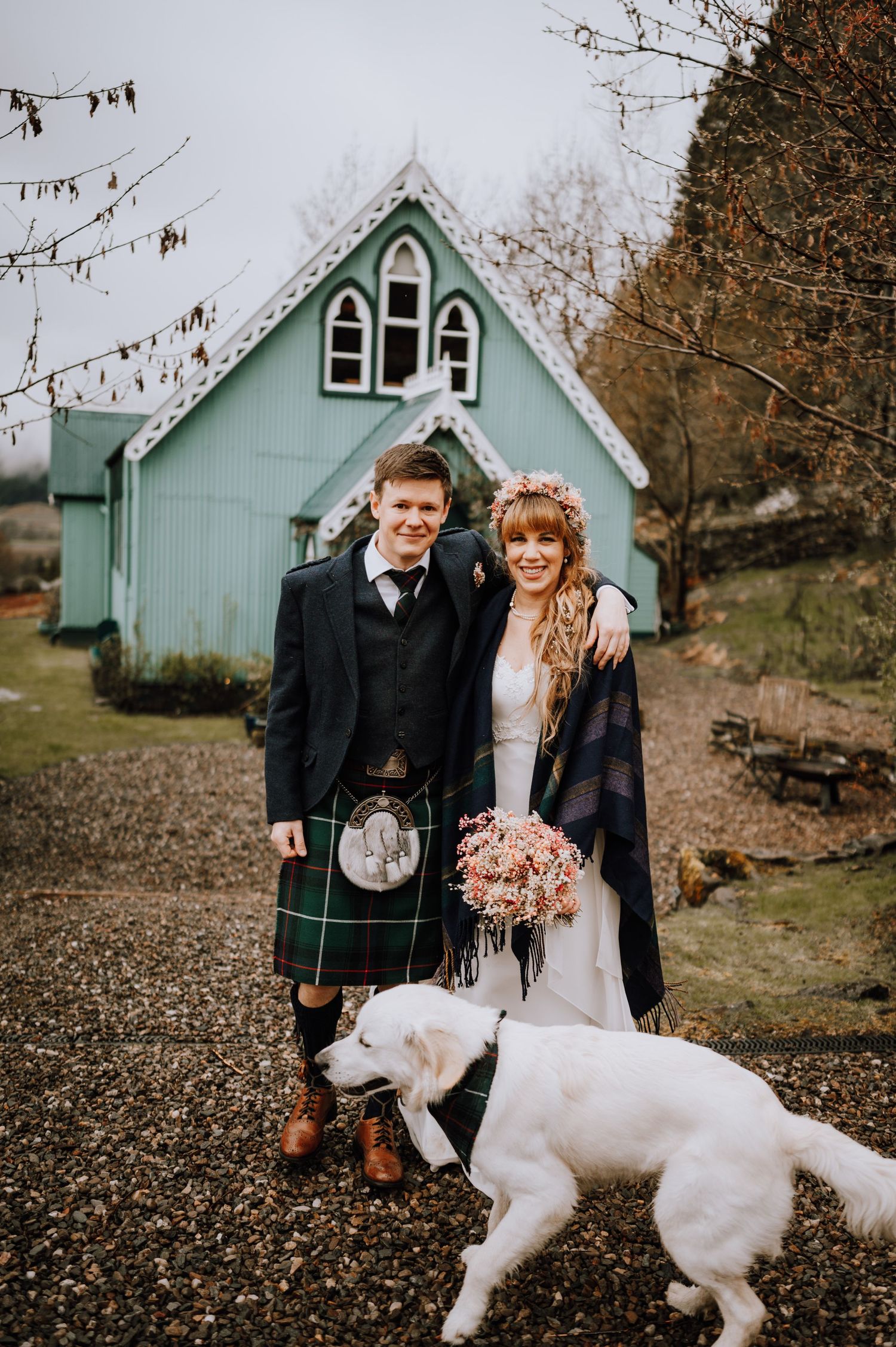 Scottish wedding couple poses with white dog outside mint-colored chapel in autumn setting.