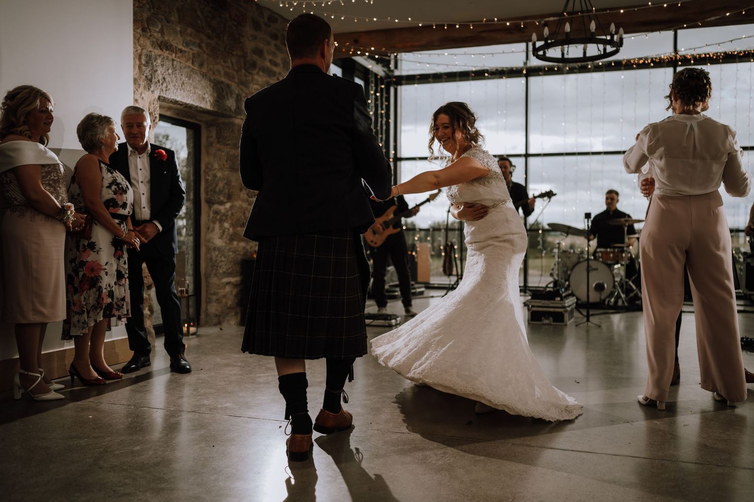 A couple dances at their wedding reception in a rustic venue with stone walls and large windows.