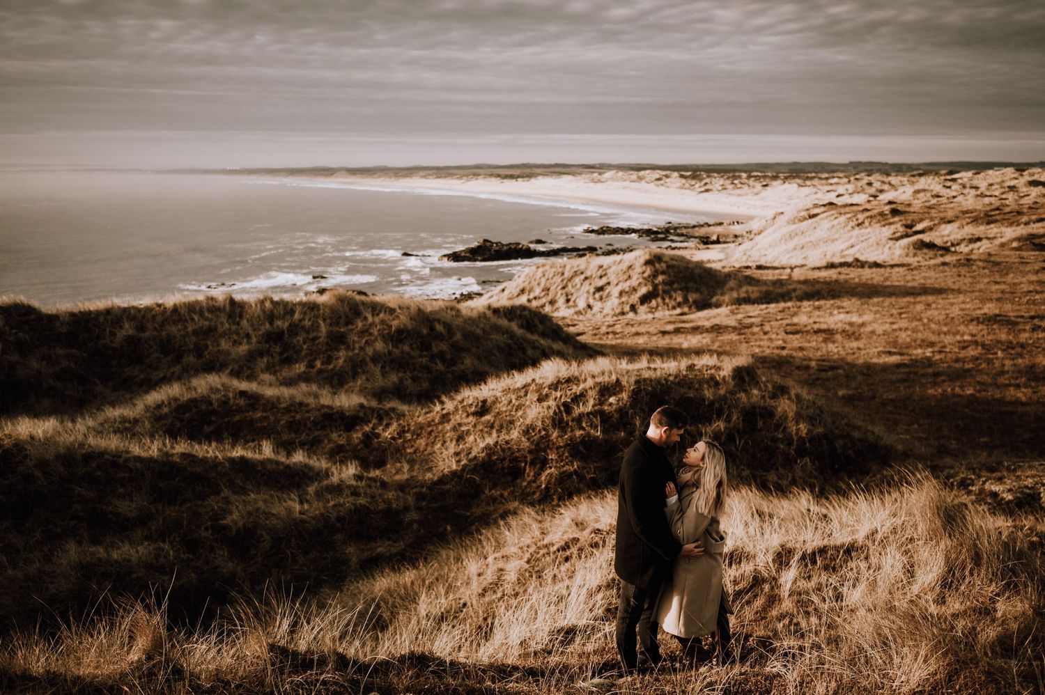 A couple embraces on coastal cliffs overlooking a dramatic seascape at golden hour.