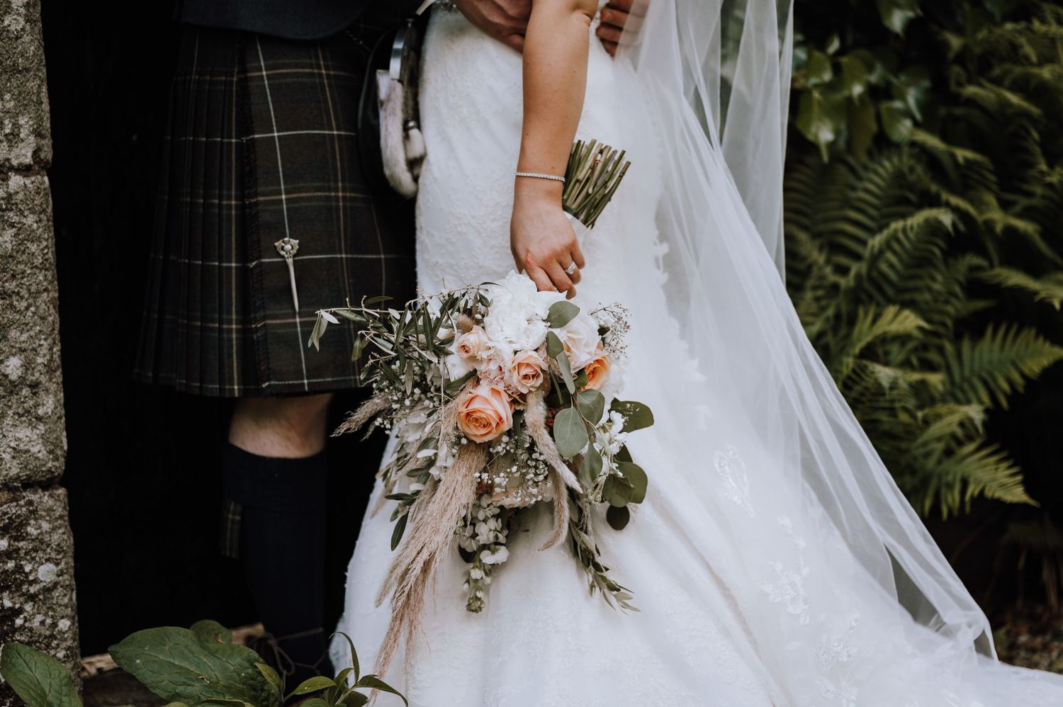 Detail shot of wedding couple with bride's bouquet featuring peach flowers and greenery.
