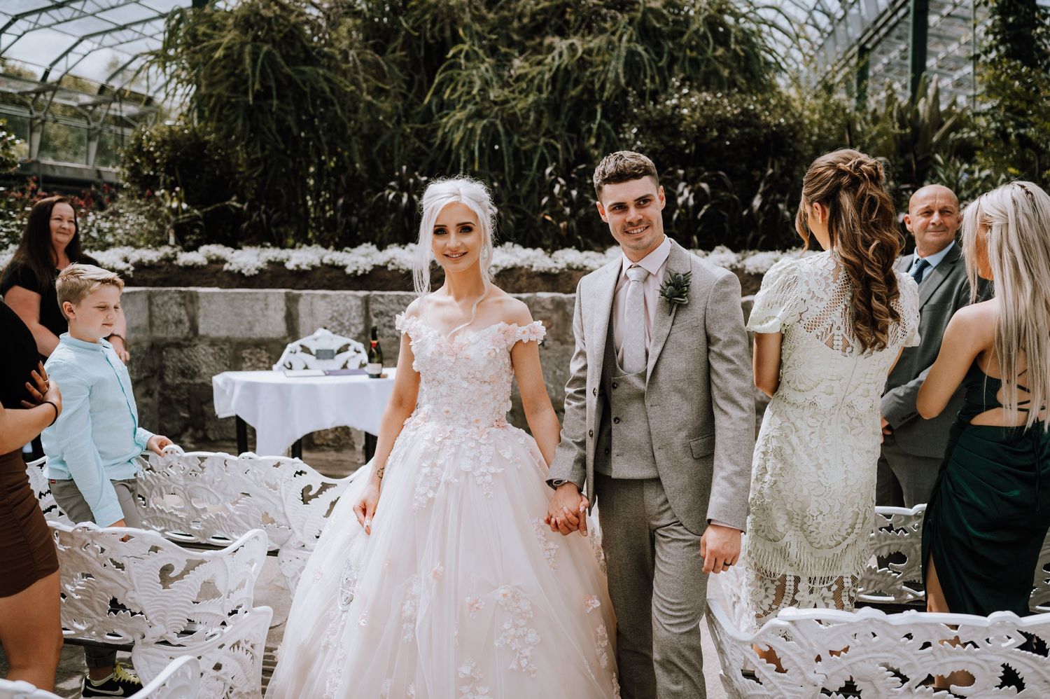 Wedding couple stands together surrounded by guests in a greenhouse with white decor.