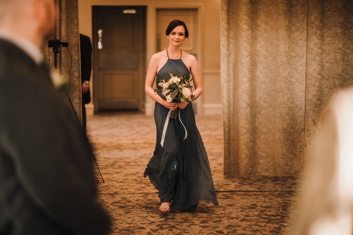 Bridesmaid in a flowing navy gown walking down a carpeted hallway holding a bouquet.