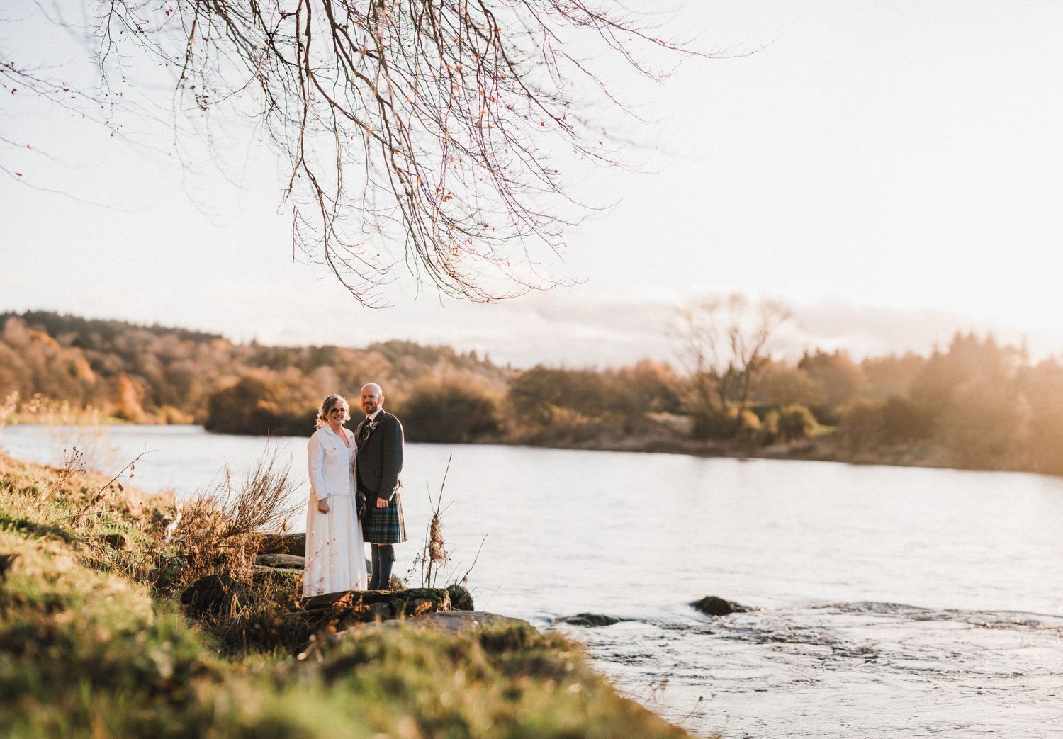 A couple stands along a riverbank at sunset with autumn trees in the background.