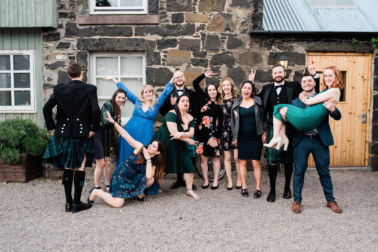 Wedding guests in formal attire pose playfully outside stone building.