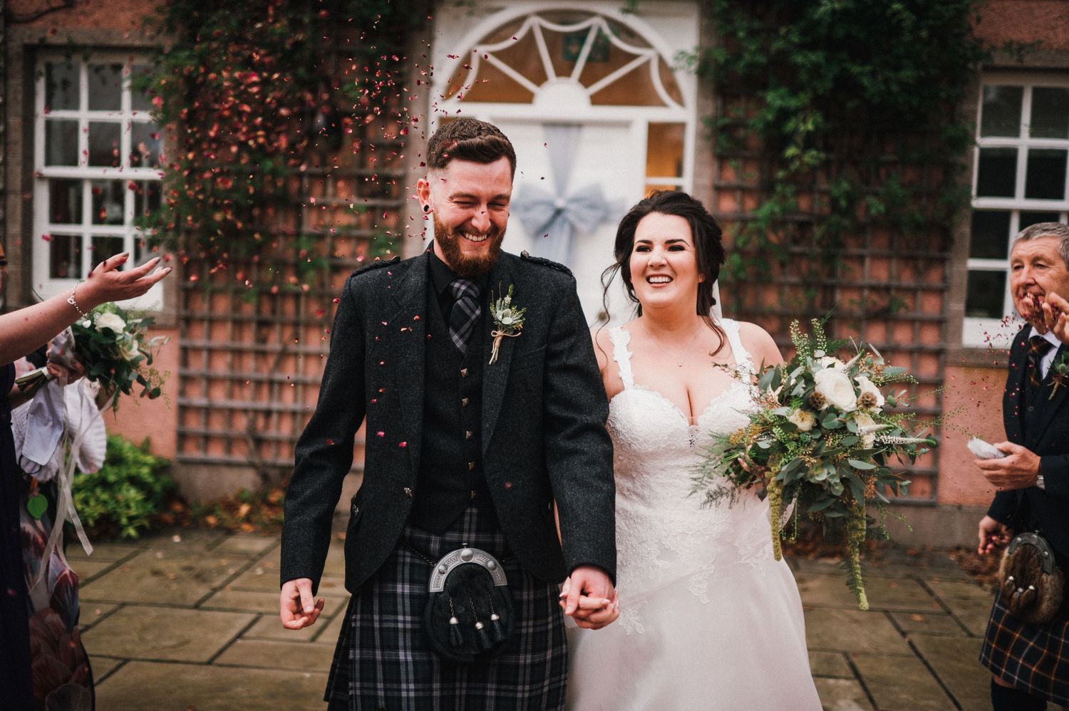 Couple exits venue in traditional Scottish and bridal attire with brick facade behind them.