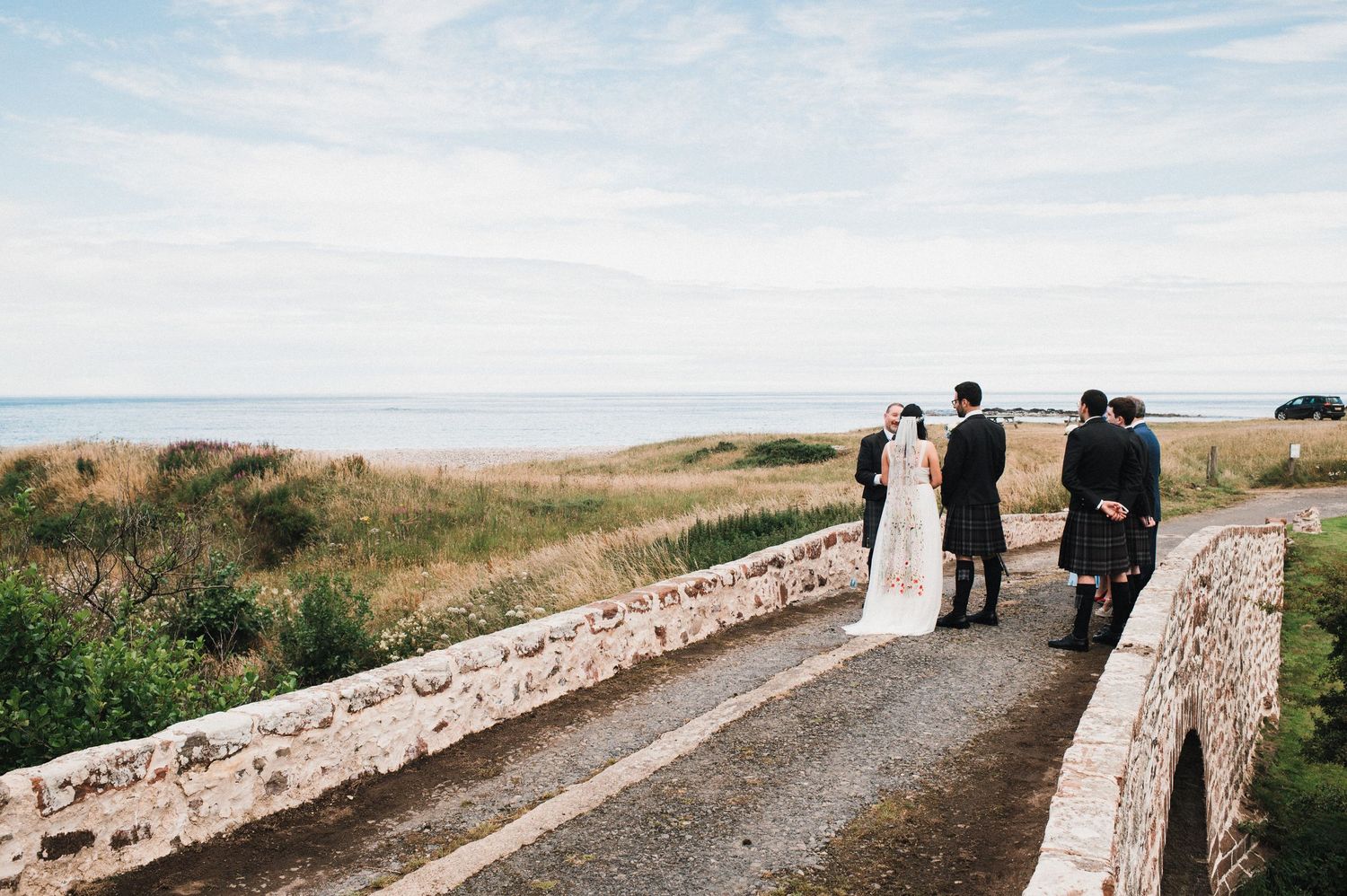 Wedding party walks along stone bridge path with coastal views.