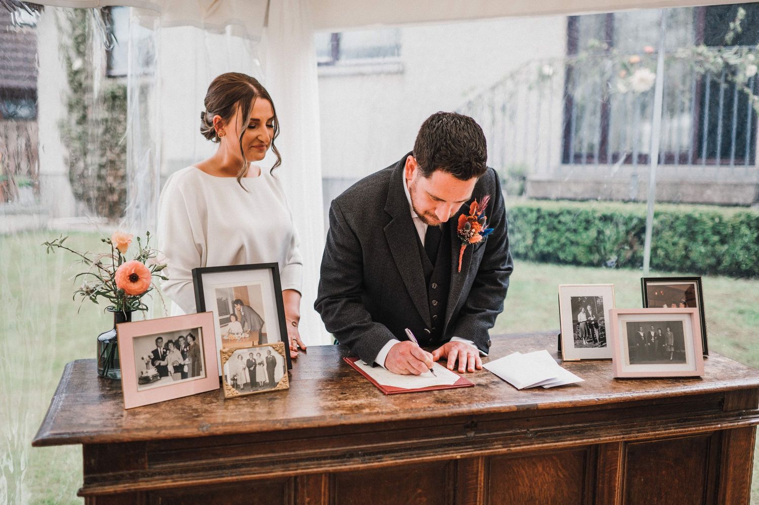 A couple signs documents at a wooden table decorated with family photos and floral arrangements at their wedding.